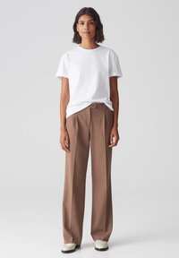 T-shirt en coton blanc à col rond avec des manches retroussées, associé à un pantalon tailleur brun taille haute. Détail de bouton remarquable à la taille.