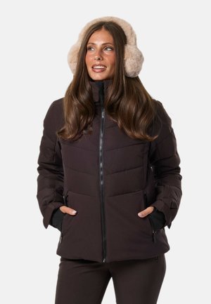 Mujer con chaqueta acolchada marrón oscuro y orejeras beige esponjosas, manos en los bolsillos de la chaqueta, mirando ligeramente de lado con un fondo neutro.