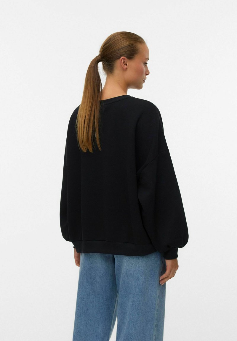 Vrouw met een lange paardenstaart, gekleed in een losse zwarte sweatshirt en wijdvallende blauwe jeans, zijwaarts gekeerd tegen een effen achtergrond.