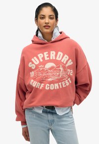 Sudadera rosa de gran tamaño con estampado gráfico en blanco que presenta olas y un surfista. Superpuesta sobre una camiseta a rayas azul y blanca, combinada con jeans claros.