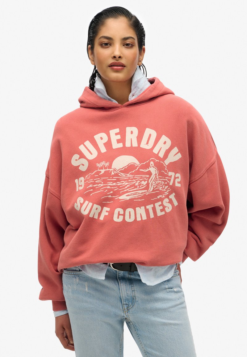 Sudadera rosa de gran tamaño con estampado gráfico en blanco que presenta olas y un surfista. Superpuesta sobre una camiseta a rayas azul y blanca, combinada con jeans claros.