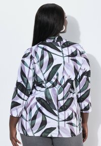 Žena s dlouhými tmavými vlasy, oblečená v bílém topu se zeleným a fialovým abstraktním listovým vzorem, stojící zády k jednoduché bílé zdi.