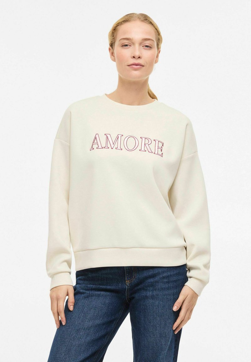 Sweat-shirt crème clair avec "AMORE" imprimé en bordeaux. Présente un col rond, des poignets côtelés et une coupe décontractée, associé à un jean foncé.