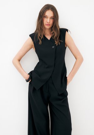 Frau mit langen braunen Haaren, die einen ärmellosen schwarzen Blazer und schwarze Hosen mit weitem Bein trägt, steht mit den Händen in den Taschen vor einer weißen Wand.