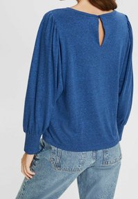 Žena v modré blůze s dlouhými rukávy a nafouknutými rukávy, se zapínáním na malé knoflíky ve vzadu nahoře, doplněné o světle modré džíny.