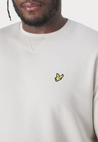Felpa grigia chiaro con scollo rotondo, polsini a coste e un piccolo logo ricamato di un uccello nero e giallo sul petto.