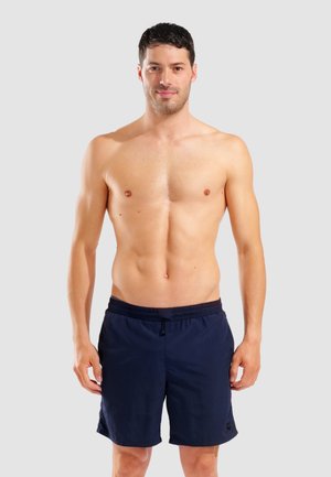 Mann mit kurzen dunklen Haaren, der marineblaue Badeshorts trägt, steht vor einem einfarbigen grauen Hintergrund und blickt entspannt nach vorne.