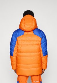 Chaqueta tipo pluma de color naranja vibrante con detalles en azul, que cuenta con capucha, textura acolchada y logo en la parte posterior. Diseño compacto e insulated.