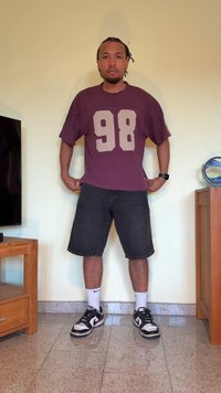 T-shirt oversized bordeaux com o número 98 em bege, combinado com calções de ganga pretos e ténis pretos. Mobiliário de madeira ao fundo.