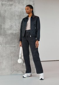 Sort bomberjakke med glidelås i front, sammen med høyhylte mørke denimjeans. Hvite sneakers og en rund, hvit crossover-veske.