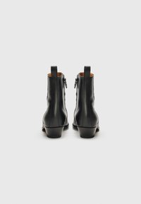 Botas de cuero negro hasta el tobillo con textura suave, tacón bajo, cremallera trasera y detalle de tira para tirar. Presentan un diseño minimalista.