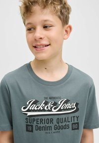 Pelēka T-kreklu, kas izgatavots no kokvilnas, ar īsām piedurknēm, ar lielu melnbaltu grafiku, kas satur uzrakstus "Jack & Jones" un "Superior Quality."