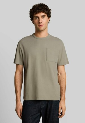 Junger Mann mit lockigem Haar, der ein hellolivgrünes, geripptes Kurzarm-T-Shirt mit Brusttasche und dunkle Hosen trägt, steht vor einem schlichten Hintergrund.