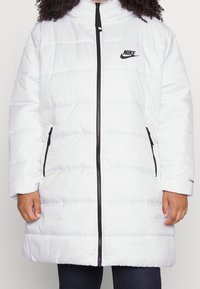 Biała kurtka puchowa z zamkiem z przodu, z poziomymi pikowanymi sekcjami i czarnym logo Nike. Posiada dwie boczne kieszenie oraz wysoki kołnierz.