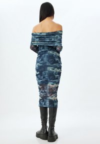 Vestido ajustado de tirantes caídos en un patrón de camuflaje azul con detalles fruncidos, combinado con botas negras altas hasta la rodilla.