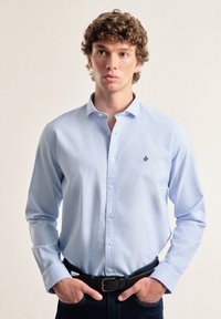 Camisa de botones azul clara con mangas largas, hecha de una tela suave, que presenta un pequeño logo negro en el lado izquierdo del pecho y un cuello clásico.