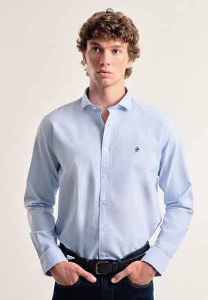 Camisa de botones azul clara con mangas largas, hecha de una tela suave, que presenta un pequeño logo negro en el lado izquierdo del pecho y un cuello clásico.