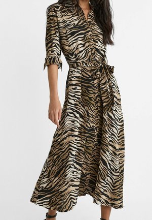Femme portant une robe chemise marron et noire à rayures tigre, taille nouée et manches retroussées, debout devant un fond uni.
