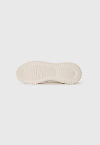 Suela de una sneaker beige de Calvin Klein con una textura en la suela y el logo grabado cerca del talón sobre un fondo blanco.