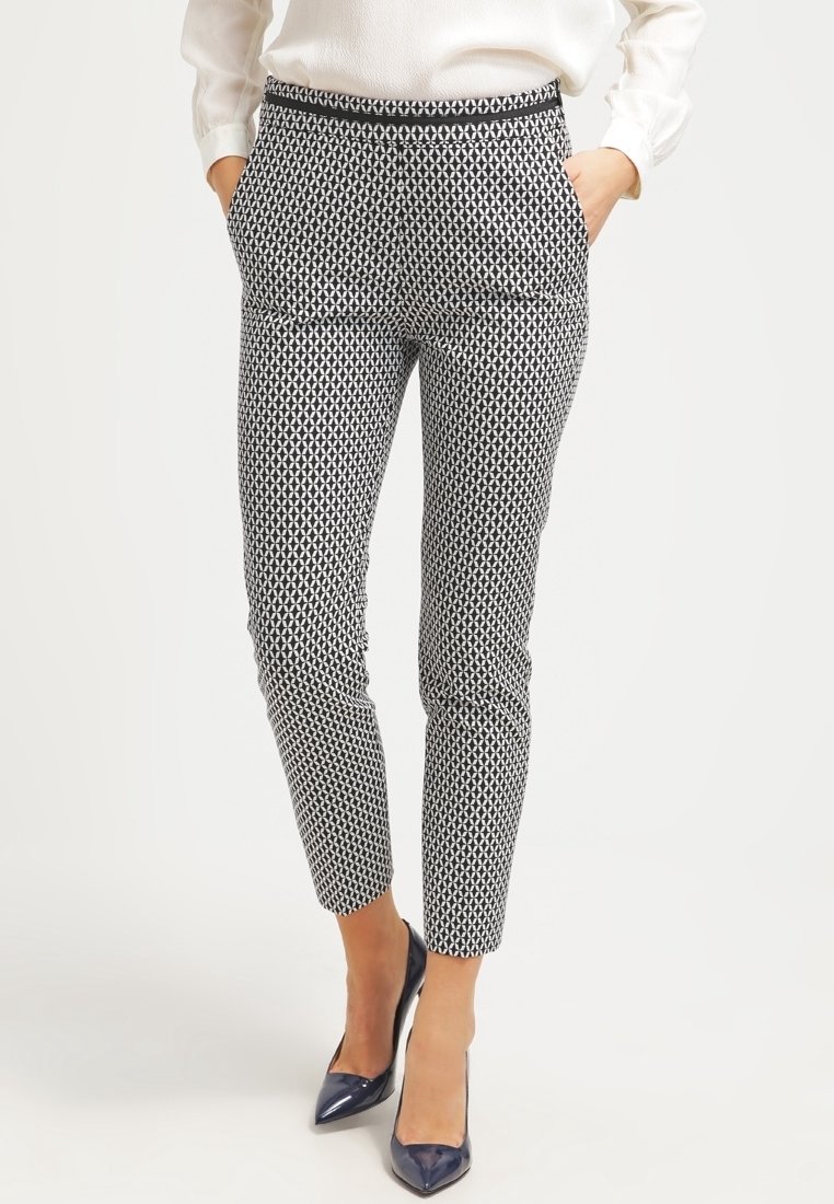 Femme portant un pantalon ankle ajusté noir et blanc à motif géométrique, une blouse blanche et des escarpins bleu marine à bout pointu.