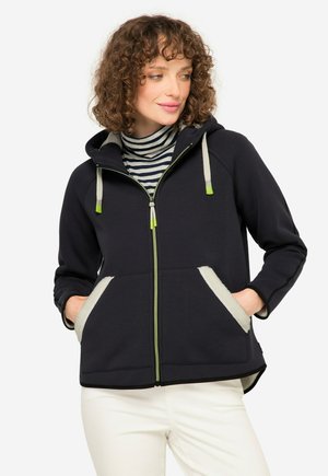 Haut à zip bleu marine avec des accents verts, poches en molleton douillet, et capuche avec cordon de serrage. Présente un col roulé rayé en dessous.