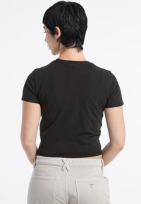 Czarny krótki t-shirt z krótkim rękawem, wykonany z miękkiej bawełny, z okrągłym dekoltem. Model nosi jasnoszare spodnie z logo na tylnej kieszeni.