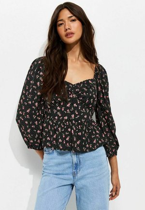 Vrouw met lang donker haar draagt een zwarte blouse met bloemmotief en pofmouwen en lichtblauwe spijkerbroek, staand tegen een effen witte achtergrond.