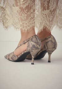 Spetsiga klackskor i ormskinnsmönster med låg klack, vristrem med rund spänne och texturerat material.