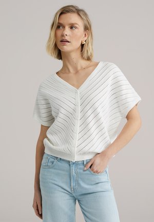 Fehér, rövid ujjú kardigán V-nyakkal, texturált, csíkos mintával és elöl gombokkal, világoskék farmernadrággal párosítva.