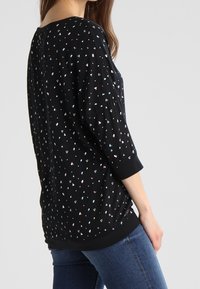 Kobieta ubrana w czarną bluzkę z małymi, wielokolorowymi serduszkami i rękawami do trzech czwarci, zestawioną z niebieskimi dżinsami, stojąca bokiem.