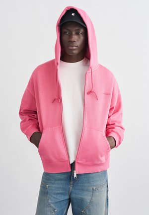 Osoba w różowej bluzie z kapturem na suwak z założonym kapturem, białej koszulce, niebieskich dżinsach i czapce czarnej, stojąca na tle jasnego, jednolitego tła.