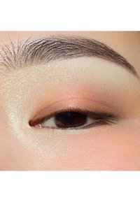 Nærbilde av et brunt øye med myk fersken- og kremøyenskygge, definert svart eyeliner og velpleid mørk bryn. Jevn hudtekstur.