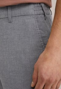 Pelēkas teksturētas bikses ar smalku pārbaudes rakstu, piedāvā elastīgu jostu un sānu kabatas. Roka atpūšas uz auduma malas.