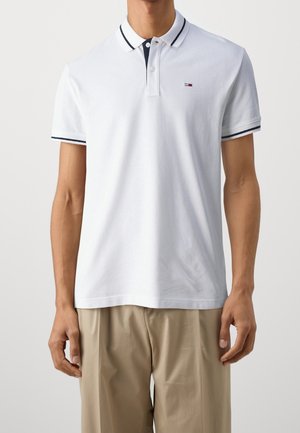 Homme portant un polo blanc avec des bordures bleu marine au col et aux manches, et un pantalon beige sur un fond uni.