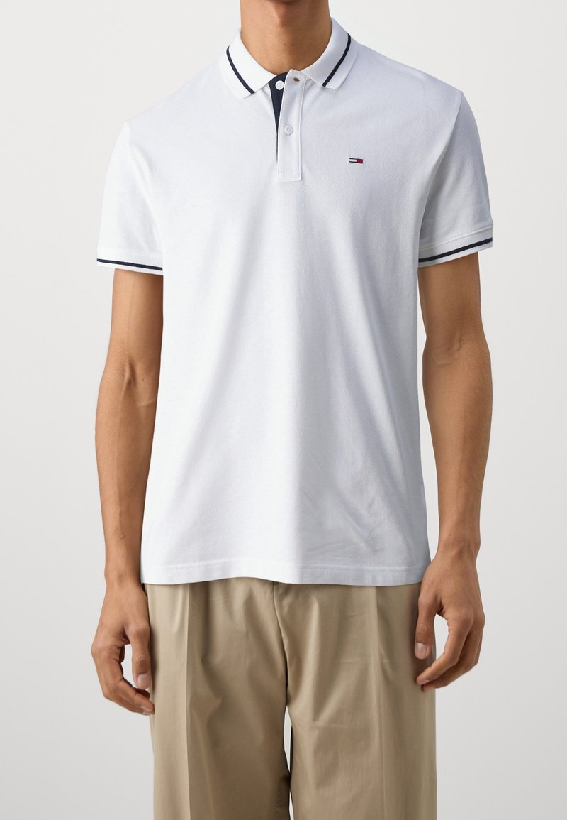 Homme portant un polo blanc avec des bordures bleu marine au col et aux manches, et un pantalon beige sur un fond uni.