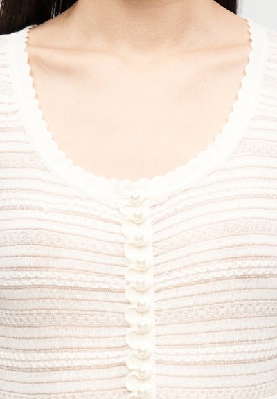 Κοντινό πλάνο ενός ατόμου που φοράει λευκό πλεκτό τοπ με υφή, με κυματιστή λαιμόκοψη και κάθετη σειρά από κουμπιά που μοιάζουν με μαργαριτάρια.