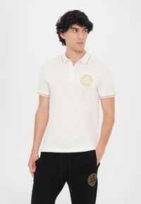 Bela polo majica z rumenimi poudarki, ki ima krožni logotip na prsih. Tkanina izgleda gladka, oblikovanje pa je klasično in prilegajoče.