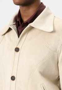 Beige suède jas met grote kraag, knoopsluiting aan de voorkant en gestikte details. Draagt over een donker bordeauxrode blouse.