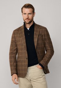 Braun karierter Blazer mit strukturiertem Finish, kombiniert mit einem dunkelblauen Pullover und hellbeigen Hosen. Der Blazer verfügt über Revers und Taschen.