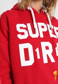 Röd hoodie i bomullsblandning med stora vita bokstäver "SUPER D1R0" över framsidan, med justerbara dragsnören och en framficka.