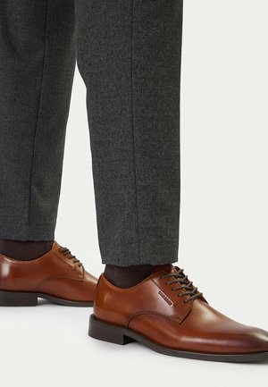 Hnědé kožené společenské boty na šněrování nošené s tmavě šedými kalhotami na míru a tmavými ponožkami na světlém pozadí.