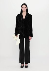 Schwarze Samt-Blazer mit klassischem Kragen und vorderen Taschen, kombiniert mit weiten schwarzen Hosen und schwarzen lackierten Ballerinas. Weißes Accessoire in der Hand.