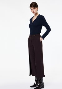 Marineblaue, gerippte Langarmbluse mit einem Kragen, kombiniert mit dunkelbraunen, hochgeschnittenen Hosen, die einen Gürtel und seitliche Schlitze haben, abgerundet mit glänzenden schwarzen Stiefeln.