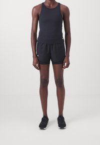 Person som står iförd svart ärmlös topp, svarta lager-på-lager idrottsshorts och svarta löparskor framför en vit bakgrund.