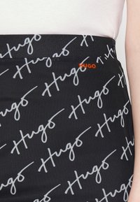 Falda negra con un patrón de script blanco de "Hugo" en toda la superficie, y un pequeño logo naranja "HUGO" en la cinturilla. Textura suave.