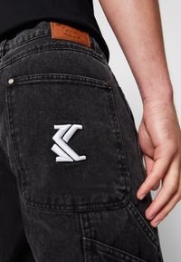 Black denim cargo pants feature a white embroidered logo on the back pocket and a brown leather patch on the waistband.