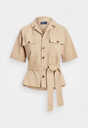 Beżowa koszula z krótkim rękawem, zapinana na guziki, z kołnierzem, dwoma przednimi kieszeniami z patkami oraz paskiem z dopasowanego materiału związanym w talii.