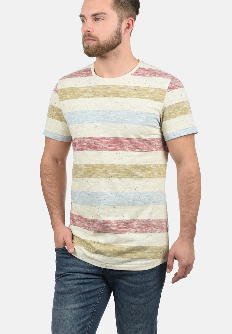 Tricou cu dungi, confecționat dintr-un material moale. Are o bază de crem deschis, cu dungi orizontale roșii, galbene și albastre, și un guler tip crew ribburat.