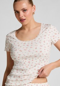 Jongeman in een witte geribbelde shirt met korte mouwen, voorzien van een klein roze bloemenpatroon en golvende randen, terwijl ze de onderkant bij haar taille vasthoudt.