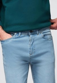 Homme portant un jean bleu clair et un pull vert foncé, une main dans la poche et l'autre ajustant l'ourlet du pull.